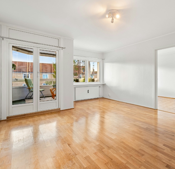 A vacant room with polished wooden floors and a spacious window, creating an airy atmosphere.